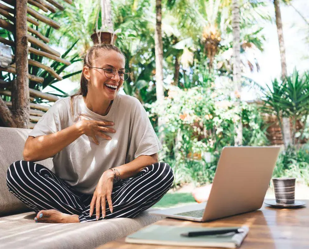 A person sitting cross-legged on a cushion in a plant-filled room, laughing, with a laptop and coffee on a low table.