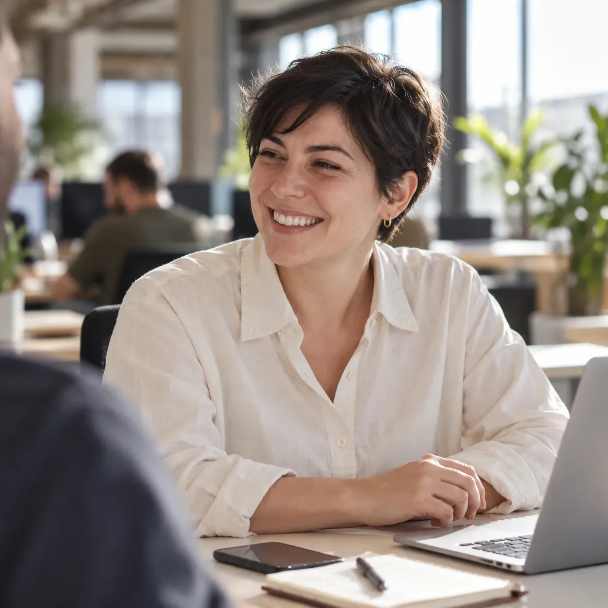 A woman with a dark pixie haircut smiling towards a colleague at a desk, with a phone, notebook and laptop nearby.