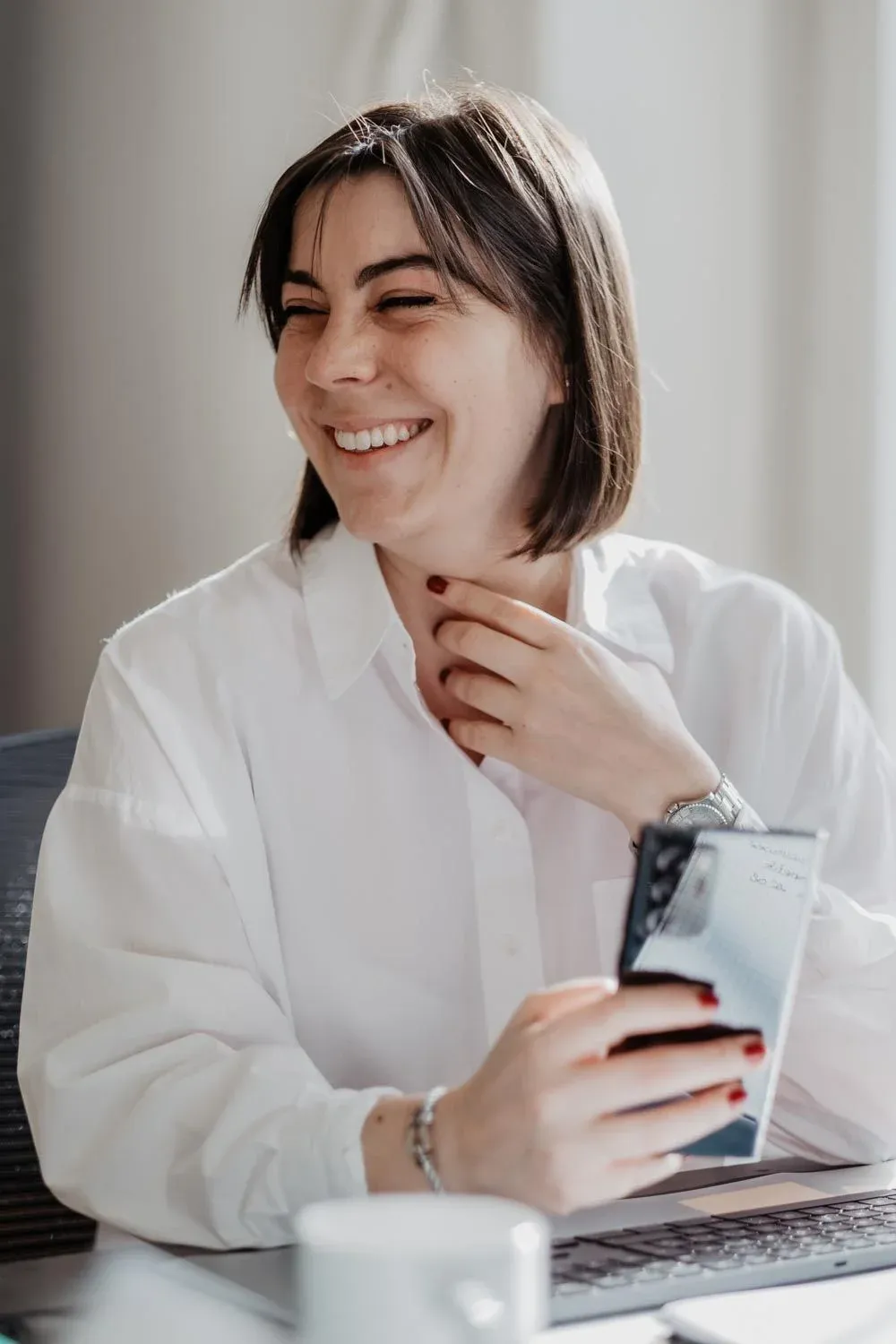 A woman laughing at her desk, holding a phone beside an open laptop.