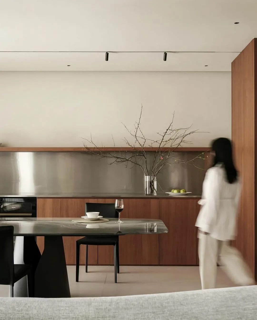 An apartment kitchen and dining area in walnut and stainless steel, with bare branches in a vase and a resident walking past.