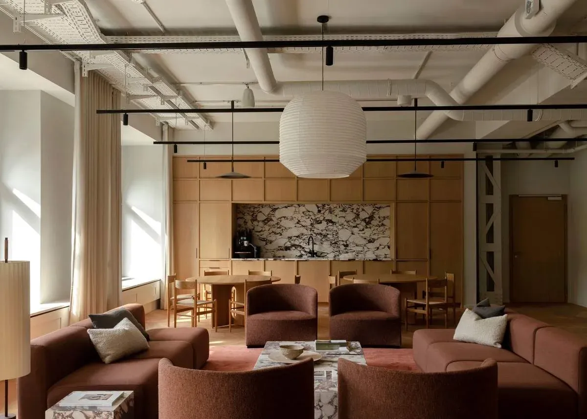 A shared co-living lounge under an exposed services ceiling, with terracotta sofas, dining tables and a marble kitchen counter behind timber cabinetry.