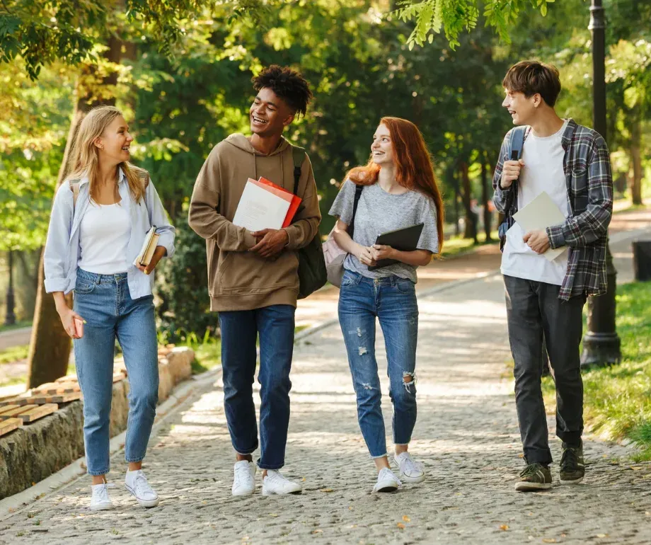 Four students walking a sunlit cobbled path through a park, carrying books and laptops.