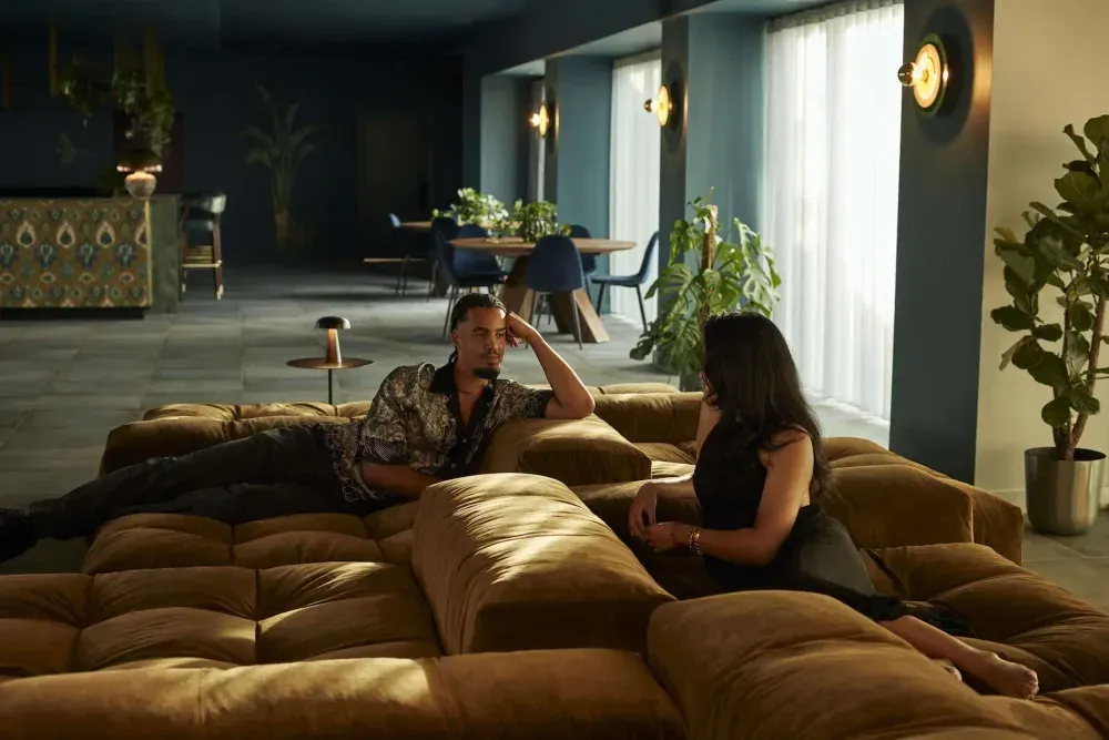 Two residents in conversation across a large tan modular sofa in a dimly lit lounge, with deep blue walls, brass wall lights and tall plants.