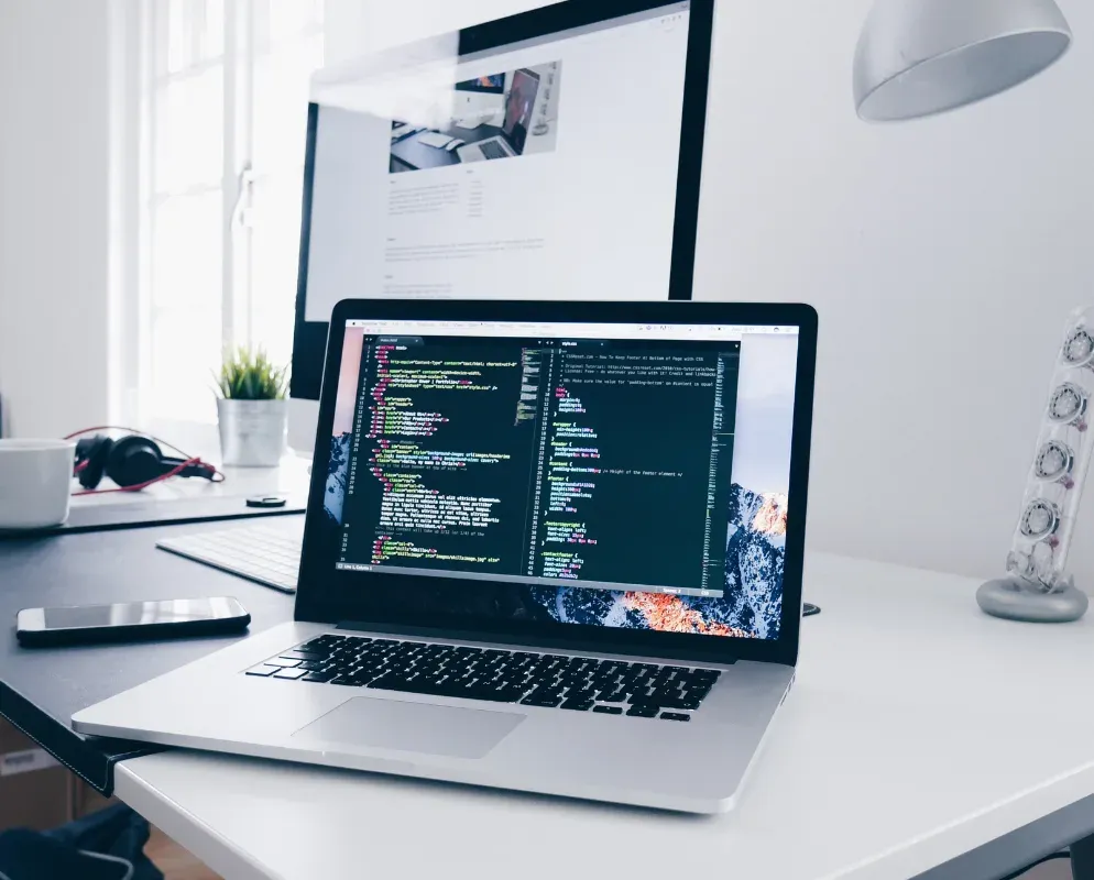 A laptop and a second monitor on a white desk, both showing code in a dark editor.