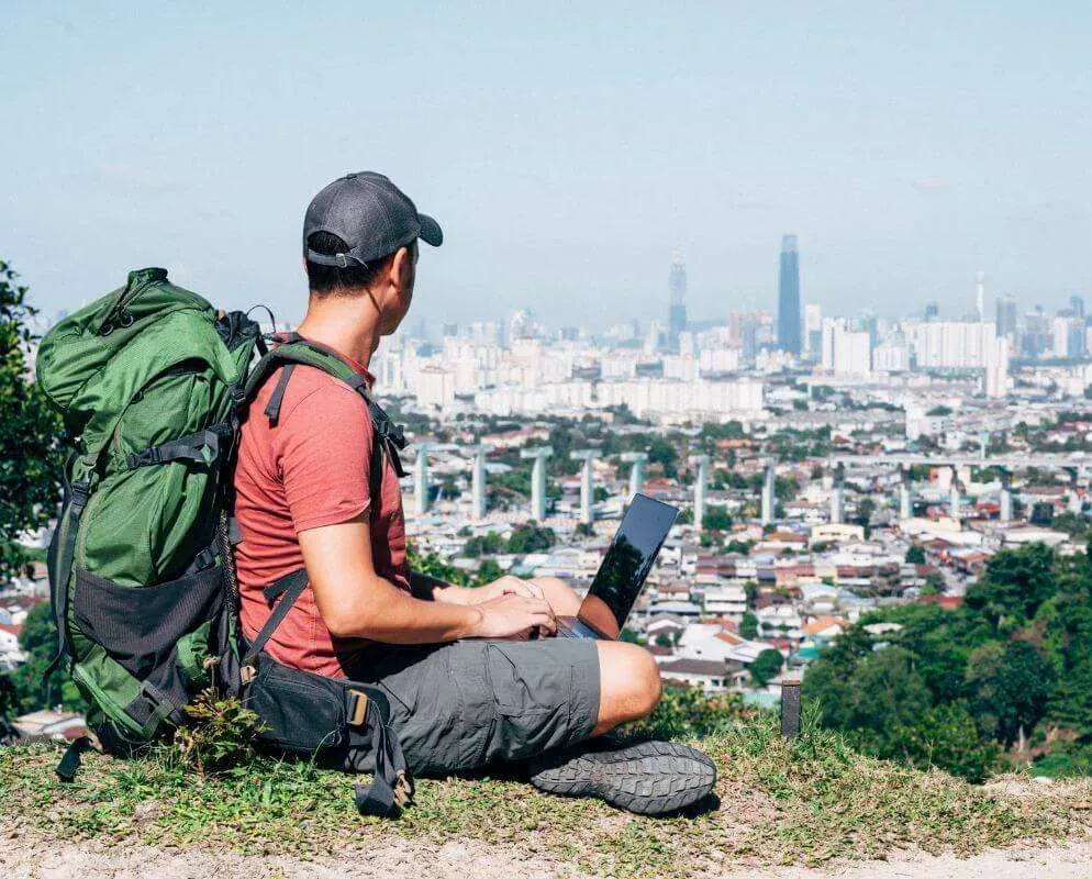 A man with a hiking backpack sitting on a hillside with a laptop, looking out over a city.