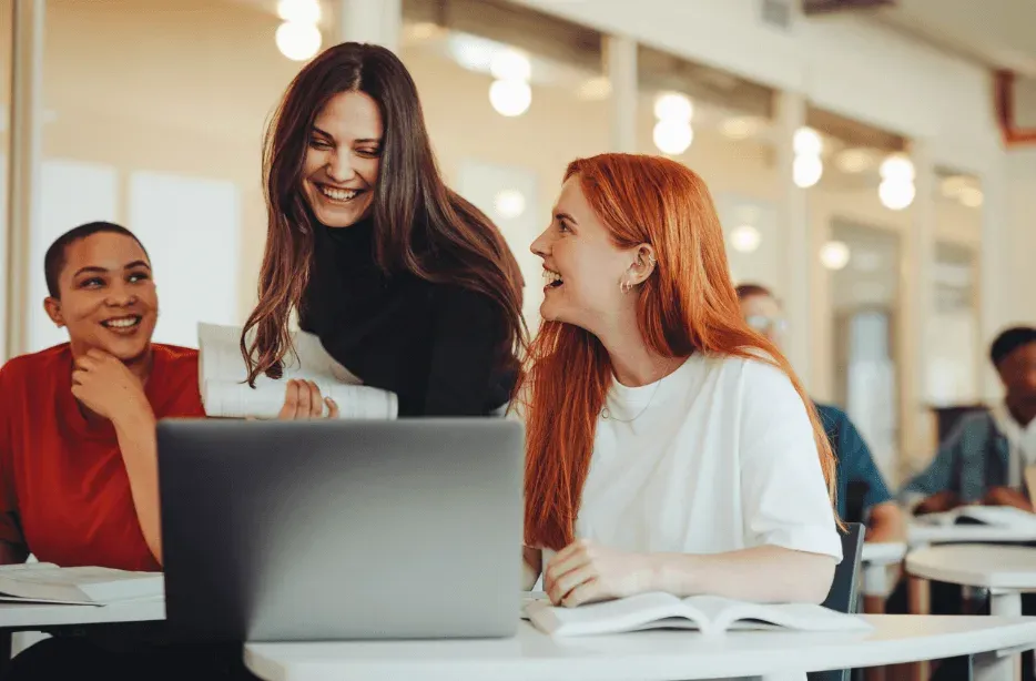 Three students laughing together over a laptop at a table in a bright shared study space.
