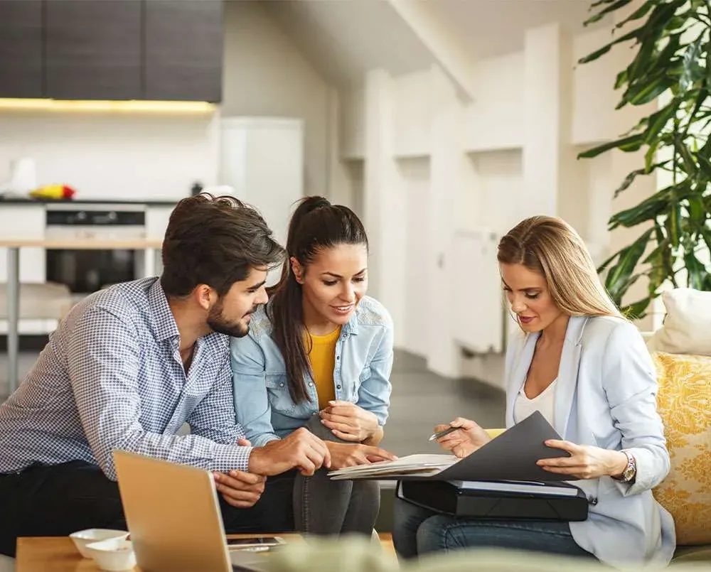Three people sitting together in a bright living room, looking at a laptop and a folder of papers.