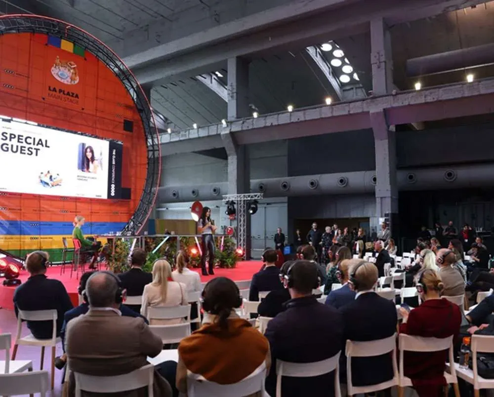 A seated conference audience facing a large circular stage screen in a high-ceilinged hall.