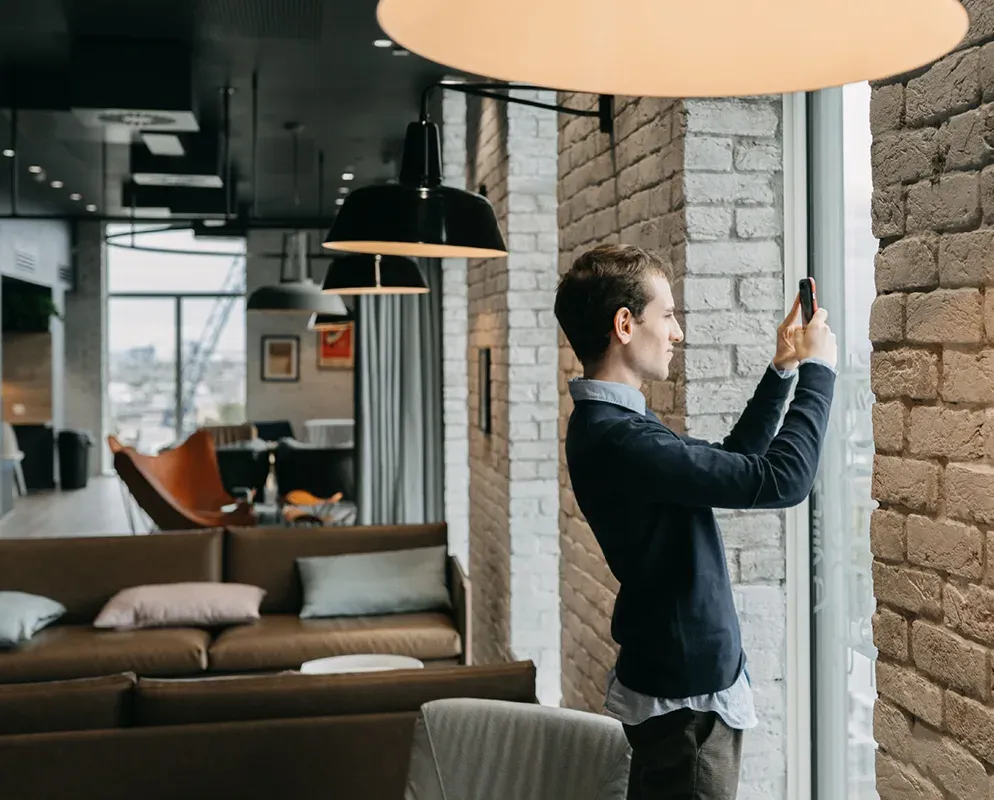 A resident photographing a warmly lit common room of exposed brick and low sofas on their phone.